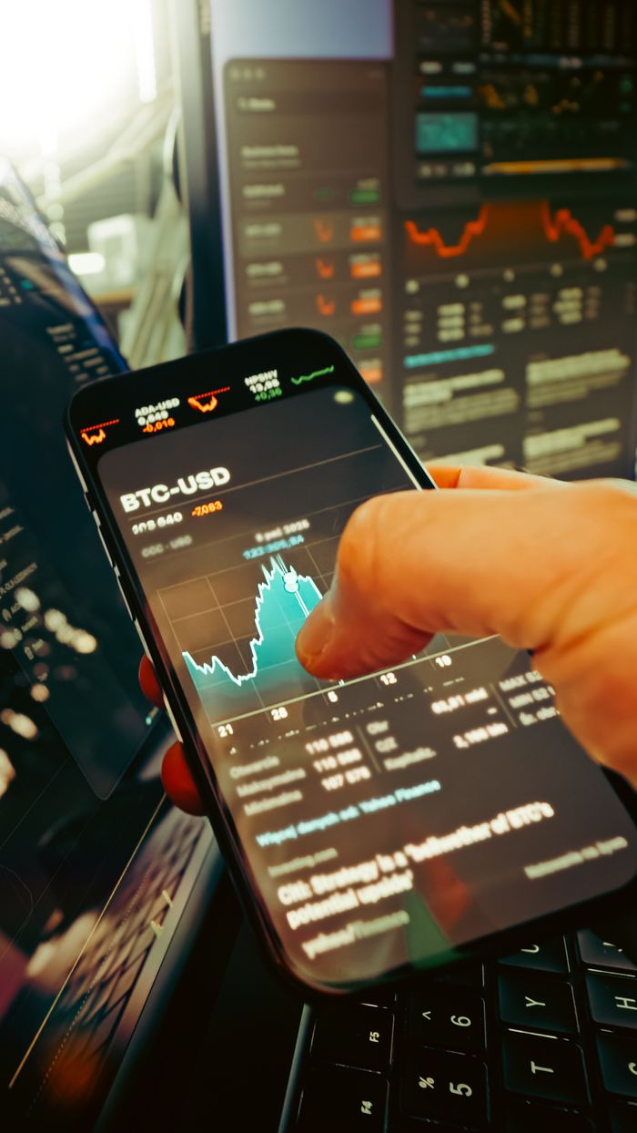 gallery-01 Une main tient un smartphone affichant un graphique BTC-USD au milieu d’écrans de trading numériques, mettant en avant la finance crypto.
